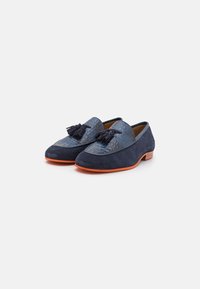 Mocasines de color azul marino que combinan cuero texturizado y ante, con borlas decorativas, suela de goma naranja y una forma redondeada elegante.