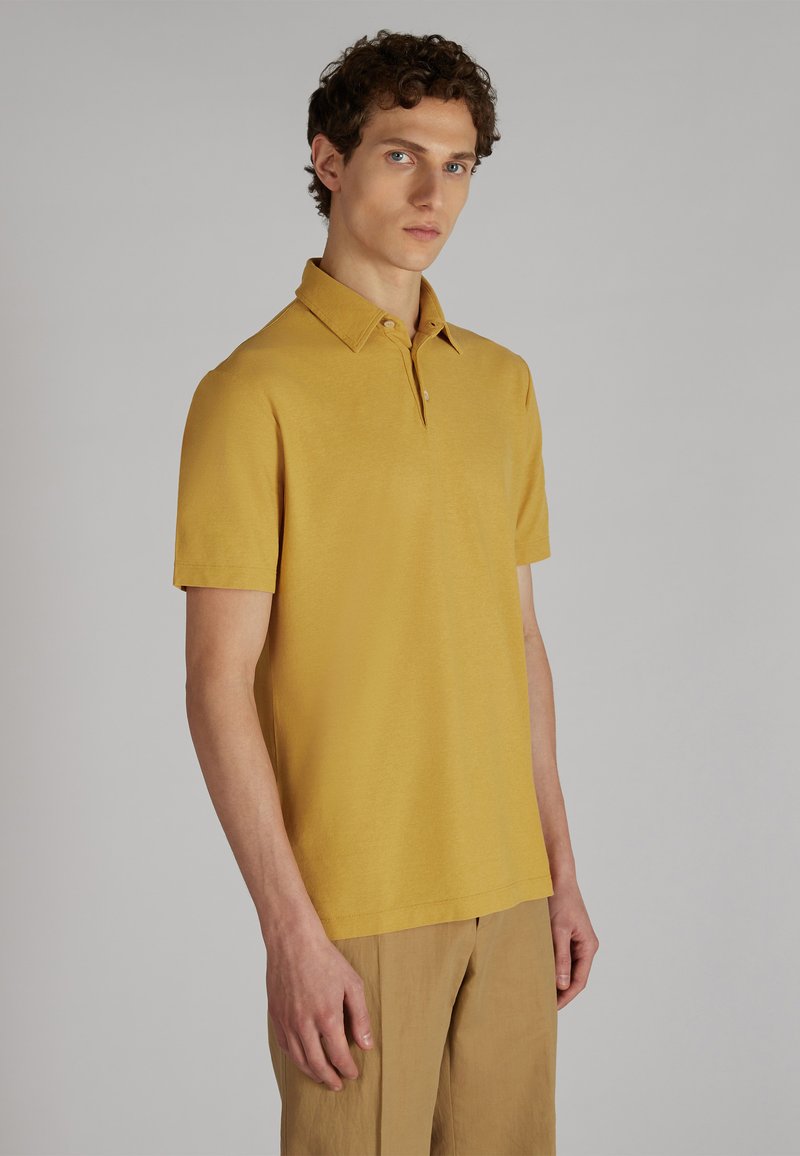 Musztardowa koszula polo z kołnierzykiem, krótkimi rękawami i kieszonką. Miękka tekstura i minimalistyczny design. Zestawiona z beżowymi spodniami.