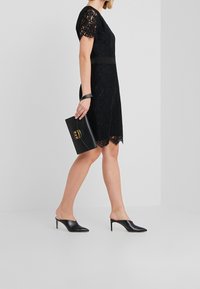 Schwarzes Spitzenkleid mit kurzen Ärmeln, tailliertem Schnitt und gewelltem Saum. Kombiniert mit einer schwarzen Leder-Clutch und offenen, hochhackigen Mules.