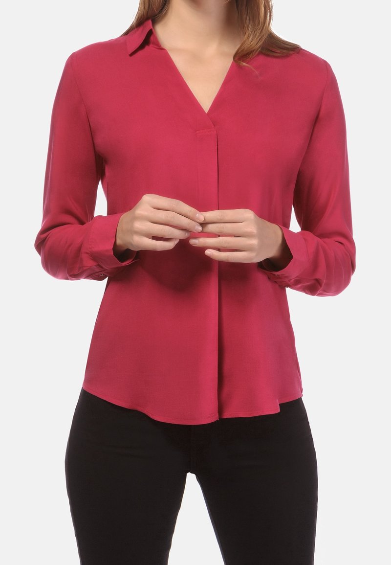 Femme portant un chemisier rouge à manches longues avec un col en V, debout les mains doucement jointes devant, portant un pantalon noir.