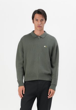Grøn strikket zip-up sweater med krave, featuring ribbede manchetter og kant, med en lille logo-patch på venstre bryst.