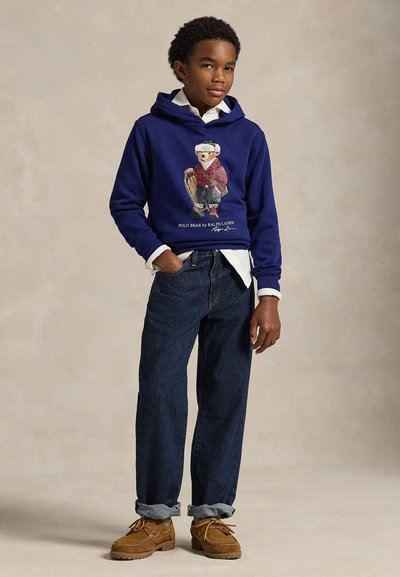 Sweatshirt azul-marinho com uma ilustração gráfica, camisa de cor clara por baixo, jeans de ganga escura e calçado bege com atacadores e textura suave.