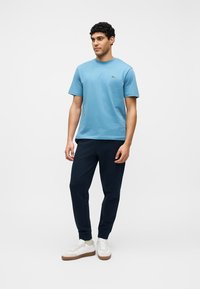 Άνδρας που στέκεται φορώντας ανοιχτό μπλε πουκάμισο με κοντά μανίκια, σκούρο μπλε παντελόνι φόρμας, λευκά αθλητικά παπούτσια και μπεζ κάλτσες σε λευκό φόντο.