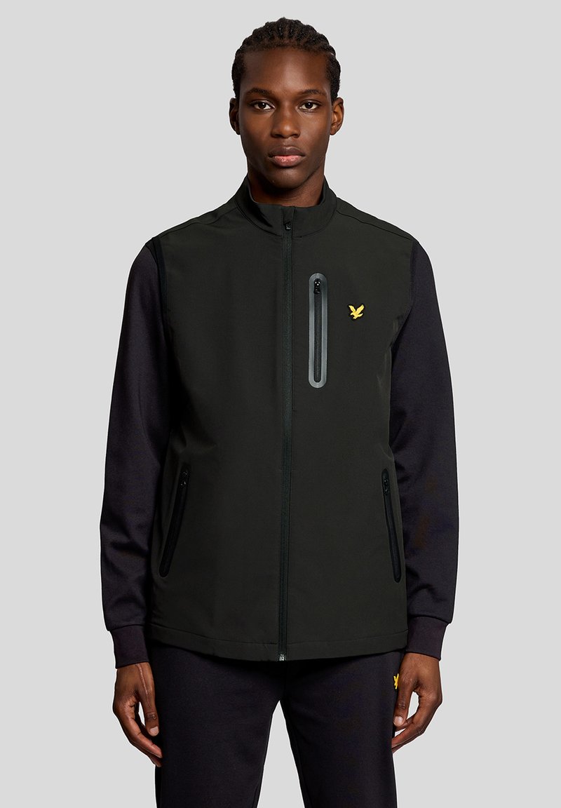 Giovane uomo con capelli intrecciati che indossa un gilet nero con zip sopra una camicia a maniche lunghe, in piedi davanti a uno sfondo grigio uniforme.