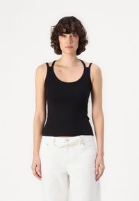 Black tank top made of soft, stretchy fabric with a rounded neckline and narrow straps. Paired with loose white trousers.