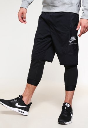 Osoba w czarnych szortach Nike na czarnych legginsach i czarnych sneakersach Nike, stojąca z jedną nogą lekko uniesioną.