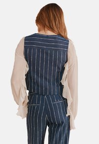 Gilet in denim con strisce bianche verticali, abbinato a maniche sheer leggere e arruffate. La combinazione di colori è blu e bianco.