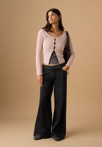 Femeie purtând un cardigan roz pal cu nasturi peste un top gri, blugi negri cu picioare largi, ținând o geantă neagră, stând în fața unui fundal bej.