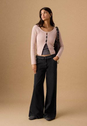 Frau trägt hellrosa Knopfstrickjacke über grauem Oberteil, schwarze Jeans mit weitem Bein, hält schwarze Handtasche und steht vor beigem Hintergrund.