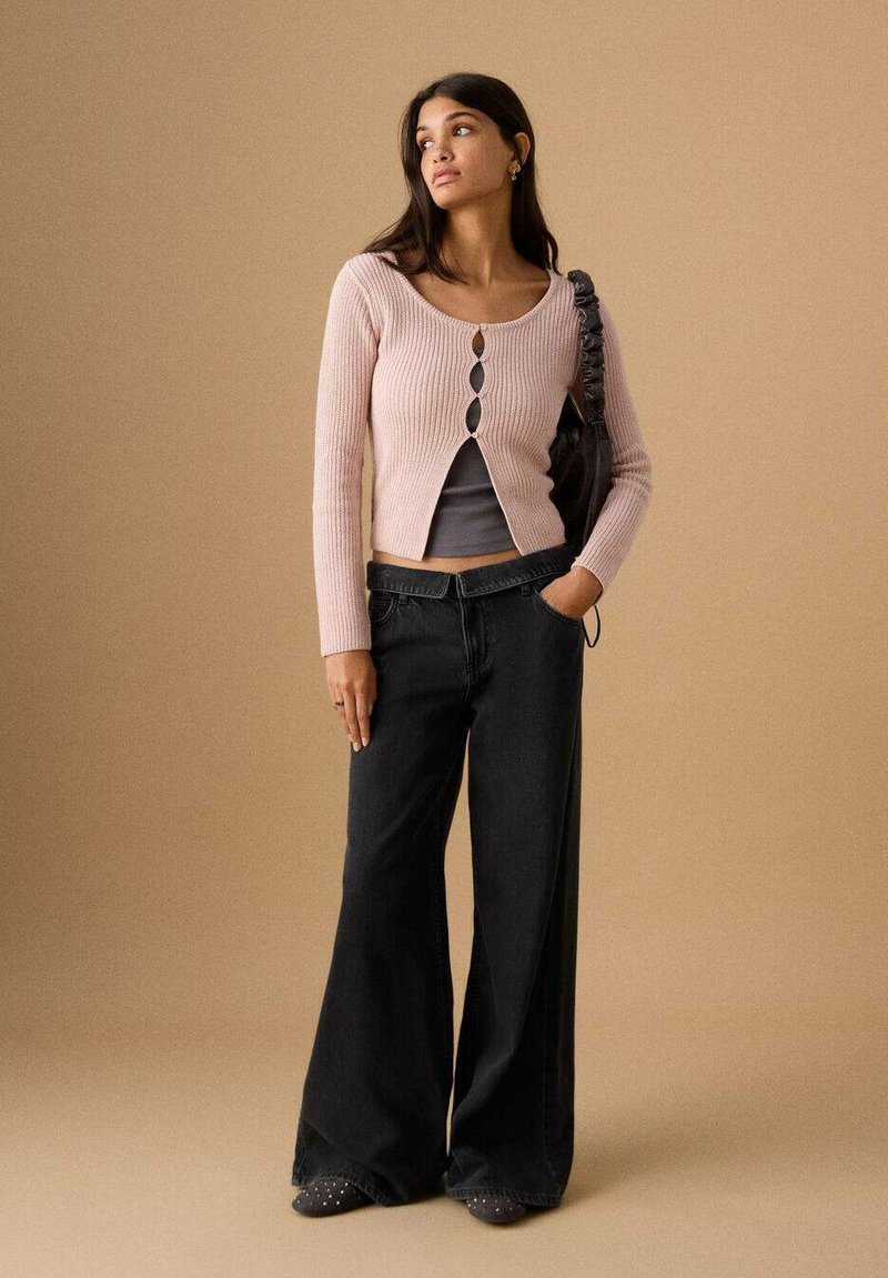 Femeie purtând un cardigan roz pal cu nasturi peste un top gri, blugi negri cu picioare largi, ținând o geantă neagră, stând în fața unui fundal bej.