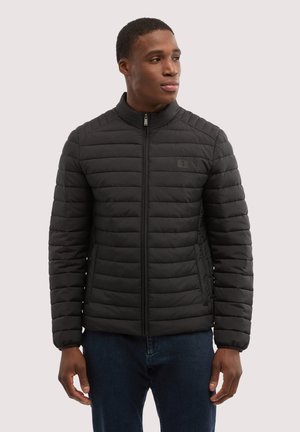 Uomo che indossa una giacca trapuntata nera con cerniera e jeans scuri, guarda verso destra su uno sfondo chiaro e uniforme.