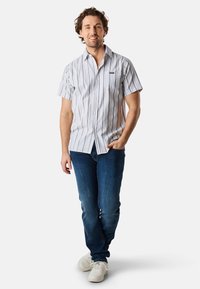 Camicia corta a maniche, con bottoni, a righe verticali blu e bianche, taschino sul petto e vestibilità rilassata, abbinata a jeans scuri e sneakers bianche.