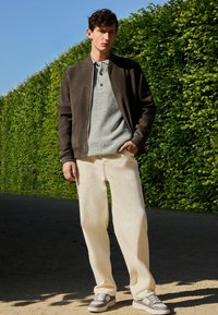 Chaqueta bomber de ante marrón sobre un polo de punto gris, combinada con pantalones de pierna ancha beige y zapatillas grises, frente a un seto verde.
