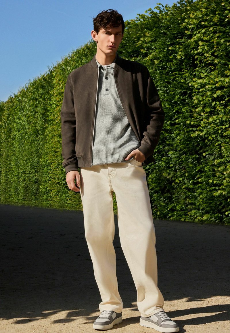 Chaqueta bomber de ante marrón sobre un polo de punto gris, combinada con pantalones de pierna ancha beige y zapatillas grises, frente a un seto verde.