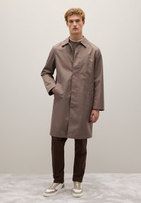 Giovane uomo in piedi con una mano in tasca, che indossa un cappotto taupe a lunghezza ginocchio, pantaloni marrone scuro e scarpe da ginnastica bianche beige.
