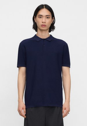 Junge Person mit glatten schwarzen Haaren, die ein dunkelblaues Polohemd mit kurzen Ärmeln trägt und vor einem schlichten hellen Hintergrund steht.