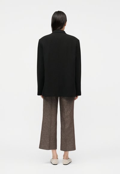 Femme debout de dos portant un blazer noir, un pantalon large marron et des chaussures beige à enfiler sur un fond blanc uni.