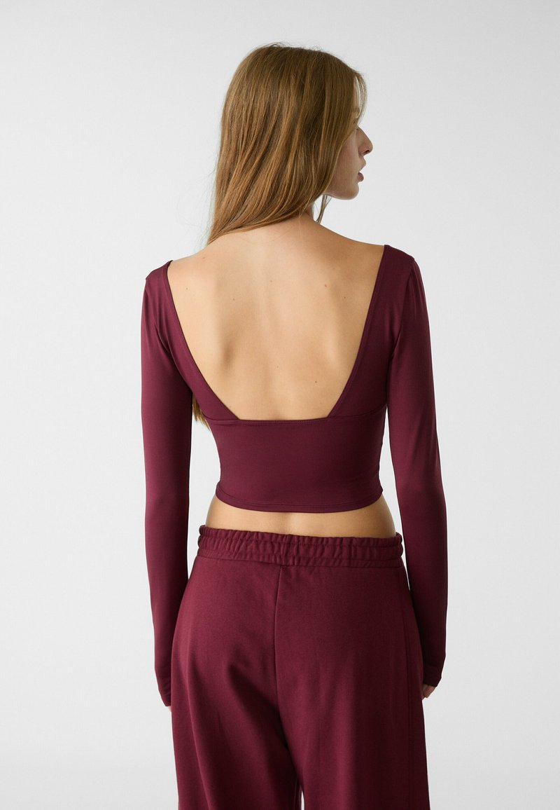 Top a maniche lunghe bordeaux, corto, con schiena a V profonda, realizzato in tessuto morbido, abbinato a pantaloni larghi coordinati con vita elastica.