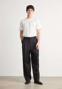 Weißes Baumwoll-T-Shirt kombiniert mit schwarzen, gefältelten Hosen. Glattes Material, lockerer Schnitt und flache schwarze Schuhe runden das Outfit ab.