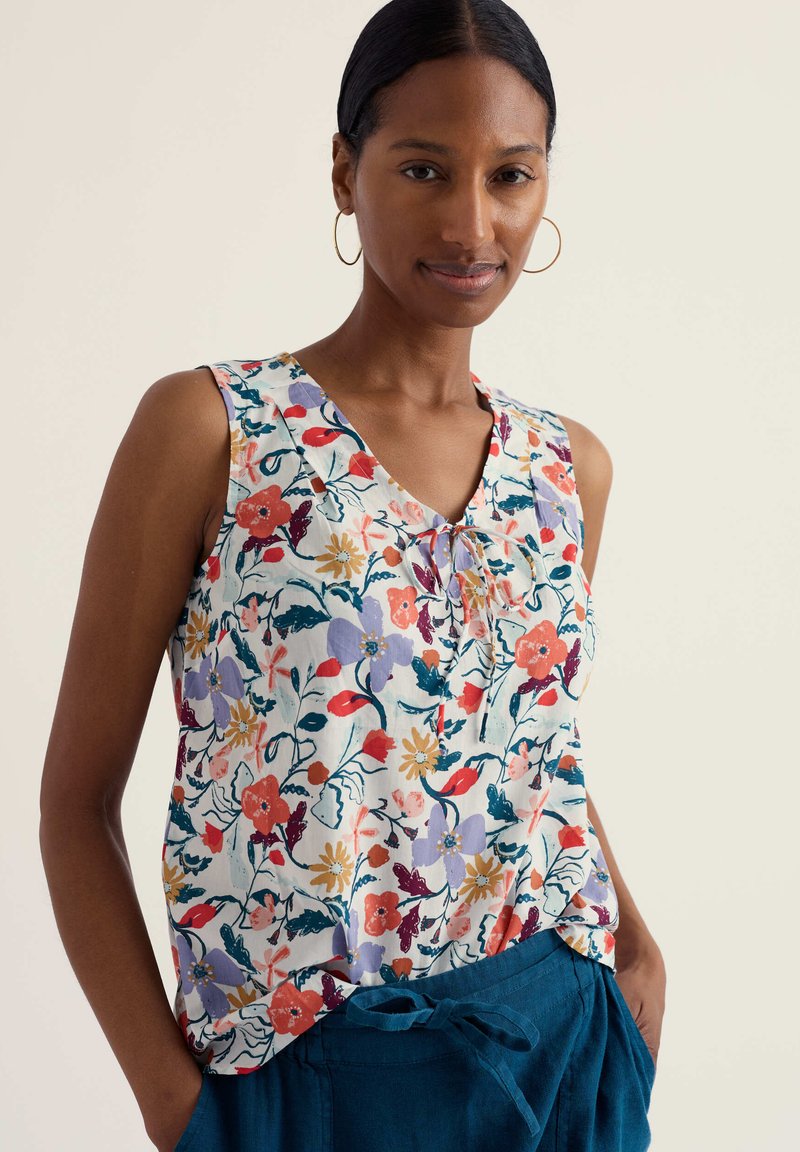 Vrouw met een mouwloze blouse met een bloemmotief in verschillende kleuren en blauwe broek met een strik in de taille, staand met handen in de zakken.