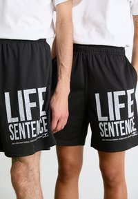 Fekete sportnadrág hálós textúrával. Mindkét oldalon nagy fehér "LIFE SENTENCE" felirat található. Rugalmas derékpánt, két látható zseb.