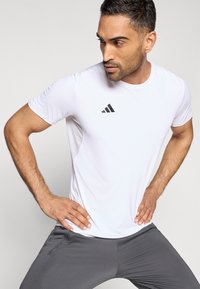 Hvid atletisk t-shirt med korte ærmer og sort adidas-logo på venstre bryst, kombineret med grå tætsiddende bukser, der demonstrerer en dynamisk positur.