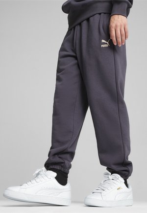 Personne portant un pantalon de survêtement Puma gris foncé et des baskets blanches Puma, debout devant un fond gris.