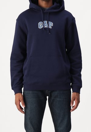 Person iført en mørkeblå GAP-hoodie med en forlommer og snore, parret med mørkeblå jeans, stående mod en hvid baggrund.