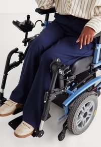 Blauwe elektrische rolstoel met zwarte zitting, marineblauwe wijde broek en beige sneakers. Focus op de details van de armbediening en het ontwerp van de wielen.