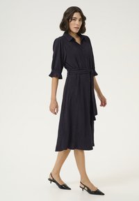 Marinblå knapp-up klänning med korta ärmar och volangmanschetter. Inkluderar ett midjebälte, texturerat tyg och en mittlång fåll. Matchas med svarta slingbacks.