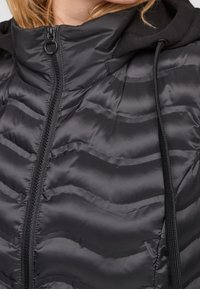 Schwarze Pufferjacke mit wellenförmigem Steppmuster, Reißverschluss und Kapuze. Verfügt über kontrastierende schwarze Kordeln und eine glatte, glänzende Textur.