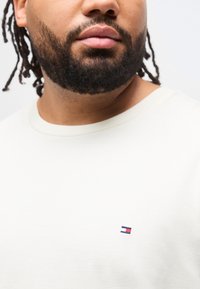 Κοντινό πλάνο ενός άνδρα με γενειάδα και πλεγμένα μαλλιά, που φοράει λευκό πουκάμισο με ένα μικρό κόκκινο, λευκό και μπλε λογότυπο στο στήθος.