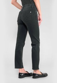Person som bär svarta slim-fit jeans och svarta lackskinnsläderskor, står mot en enkel ljus bakgrund.