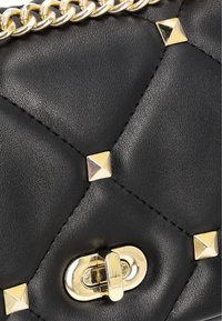 Borsa a tracolla in pelle nera trapuntata con borchie a forma di piramide color oro e una chiusura rotonda. Accentuata da una spessa tracolla in catena dorata.