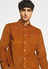 Camisa de pana marrón con cuello abotonado, bolsillo en el pecho y pequeño logo bordado en el bolsillo. Presenta un tejido texturizado y cierre con botones.