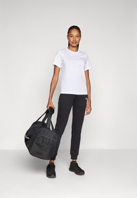 Witte T-shirt met een klein logo, zwarte trainingsbroek met enkelboordjes, en een zwarte sporttas met een tekstaccent. Zwarte sportschoenen.