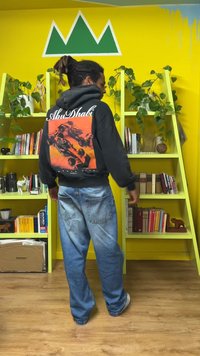 Sweat à capuche noir avec un imprimé graphique de voitures de course et le texte "AbuDhabi". Jean bleu ample avec une texture délavée. Bibliothèque intérieure et plantes en arrière-plan.