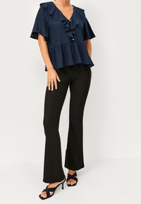 Kvinna klädd i en marinblå volangblus med kort ärm, svarta slim-fit flarebyxor och svarta klacksandaler med korsade remmar.