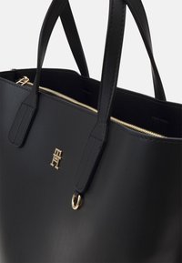 Borsa a spalla in pelle nera con hardware dorato, dotata di manici doppi, chiusura con zip e logo embossato sul davanti. Texture liscia.