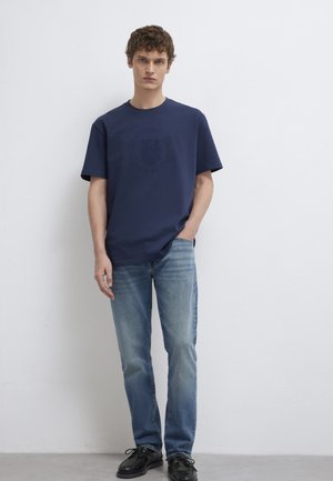 Junger Mann mit lockigem Haar, der ein dunkelblaues Grafik-T-Shirt, hellblaue Jeans und schwarze Schuhe trägt, steht mit einer Hand in der Tasche vor einer weißen Wand.