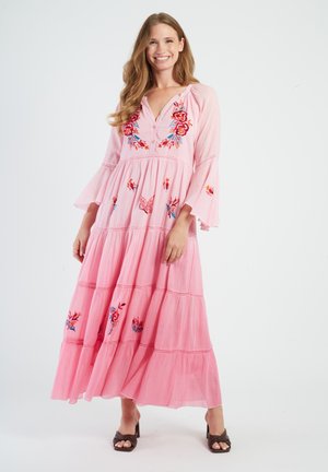 Uśmiechnięta kobieta, ubrana w różową maxi sukienkę z kwiatowym haftem, dzwonkowatymi rękawami i brązowe, odkryte szpilki, stoi na białym tle.