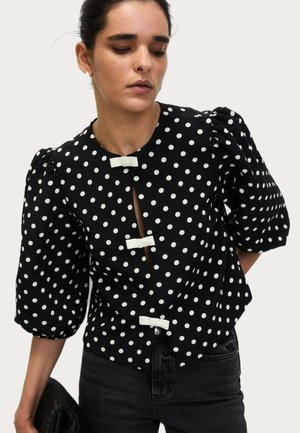 Schwarze Bluse mit weißen Polka-Dots, gepufften Ärmeln und einem Schlüsselloch-Ausschnitt, mit dekorativen weißen Schleifenverschlüssen an der Vorderseite.