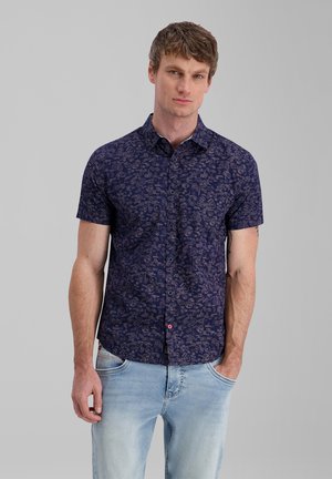 Mann trägt ein kurzärmeliges dunkelblaues Hemd mit Knöpfen und hellem Muster sowie hellblaue Jeans, steht vor einem einfarbigen Hintergrund.