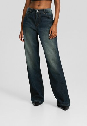 Femme portant un jean noir délavé à jambes larges et des chaussures noires à bouts pointus, debout devant un fond gris uni.