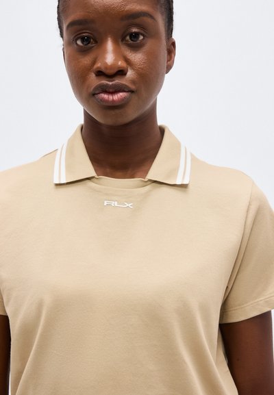 Νεαρή γυναίκα που φοράει μπεζ πόλο μπλούζα με λευκές ρίγες στο γιακά και το λογότυπο "RLX" στο στήθος, πάνω σε απλό φόντο.