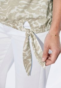 Lichtgewicht, beige top met dierenprint en geplooide zoom. Strikken zijn aan de zijkant vastgemaakt, gecombineerd met witte broek. Soepele textuur.