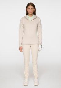 Felpa con zip beige e cappuccio, abbinata a pantaloni beige aderenti. Presenta una camicia colletto verde chiaro sotto e un guanto da golf bianco.