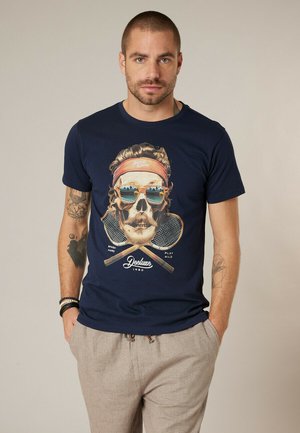 Homme aux cheveux courts et tatouages, portant une chemise bleue marine avec un motif de crâne portant des lunettes de soleil et un bandeau, des raquettes de tennis croisées, et un pantalon beige.