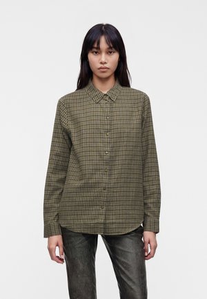 Femme portant une chemise à carreaux verte boutonnée et un pantalon foncé, debout devant un fond clair uni, regardant droit devant.