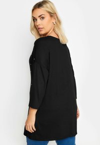 Musta pitkähihainen paita, jossa on rento istuvuus, pyöreä pääntie ja koristeelliset hopeiset koristeet olkapäillä.